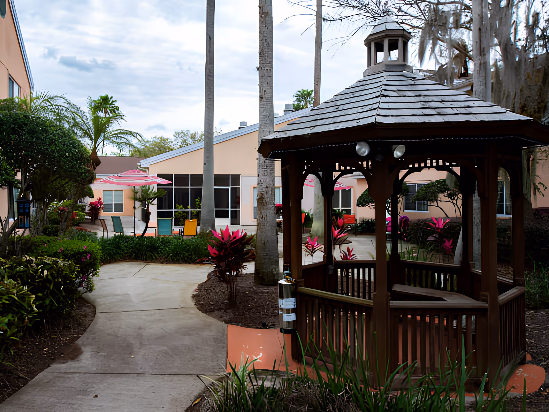 A wooden gazebo sits in a landscaped courtyard with a paved walkway, tropical plants, and patio umbrellas in front of a peach-colored building.