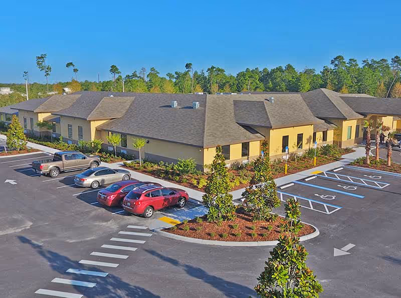 Exterior view of a single-story assisted living facility building with a large parking lot in front. Several cars are parked, including in designated handicapped spaces. The building is surrounded by landscaped greenery and trees, with a clear blue sky overhead.