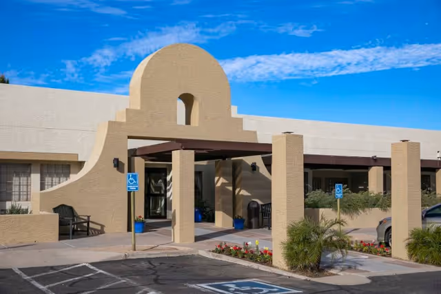 Entrance of a single-story stucco building with an arched portico, accessible parking spaces and planters under a blue sky.
