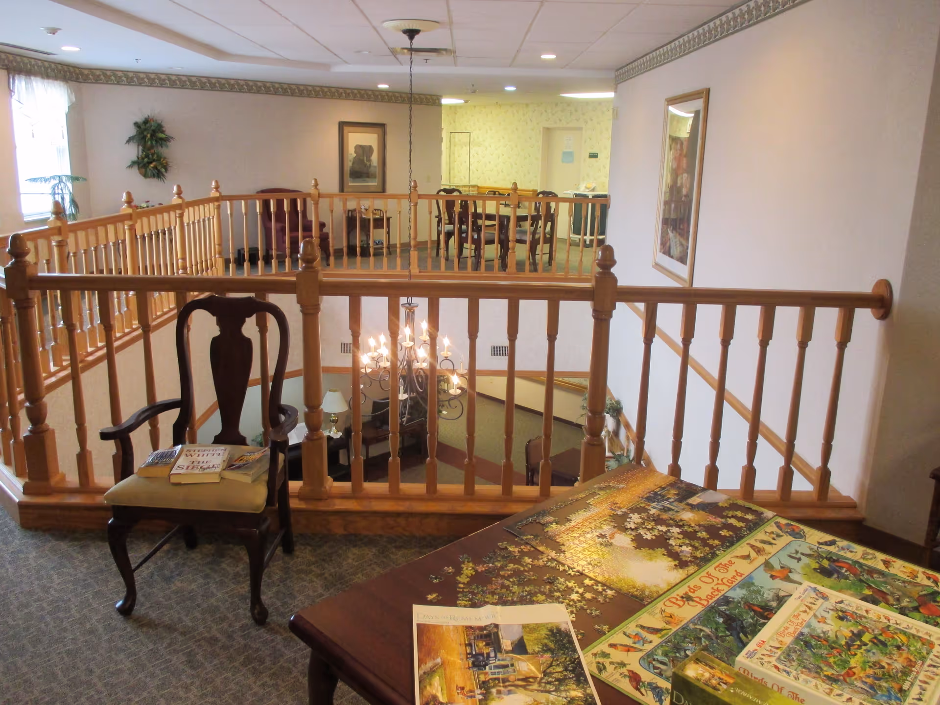Interior view of a senior living facility showing a carpeted upper floor area with wooden railing overlooking a lower level. There is a wooden chair with a cushion and some books on it, a table with an unfinished jigsaw puzzle and puzzle boxes, and a dining area with a table and chairs in the background. The walls are decorated with framed pictures and there is a chandelier hanging over the lower level.
