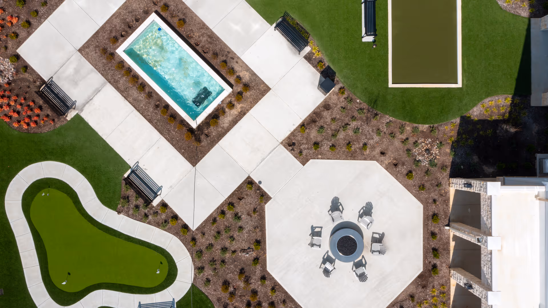 Aerial view of an outdoor recreational area featuring a small rectangular pool, a putting green, several benches, a fire pit surrounded by chairs on a concrete patio, and landscaped garden beds with pathways.
