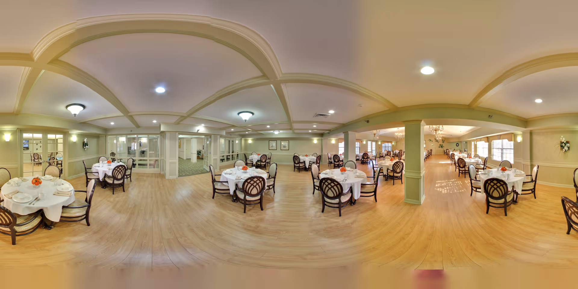 A spacious and well-lit dining room in a senior living facility with multiple round tables covered with white tablecloths, each set with plates, napkins, and small floral centerpieces. The room features wooden flooring, soft yellow walls, ceiling lights, and large windows allowing natural light to fill the space.