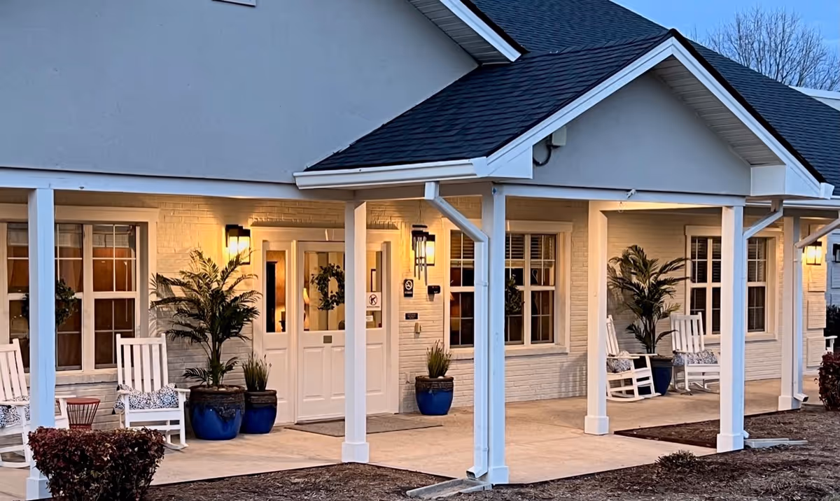 Front entrance of a senior living facility with a covered porch, white rocking chairs, potted plants, and warm exterior lighting.