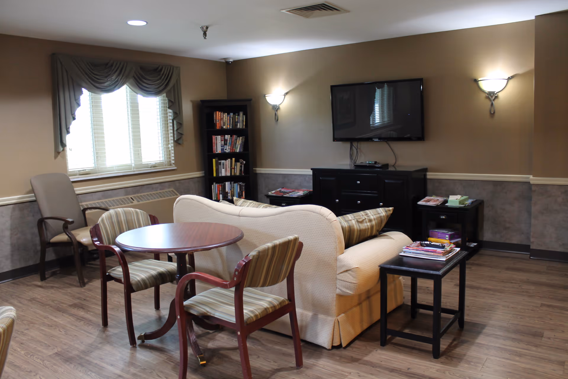 A cozy living room area with a beige couch, two striped chairs around a round wooden table, a tall bookshelf filled with books, a wall-mounted flat screen TV, and two wall sconces providing light. The room has wood flooring, a window with draped curtains, and a small side table with magazines.