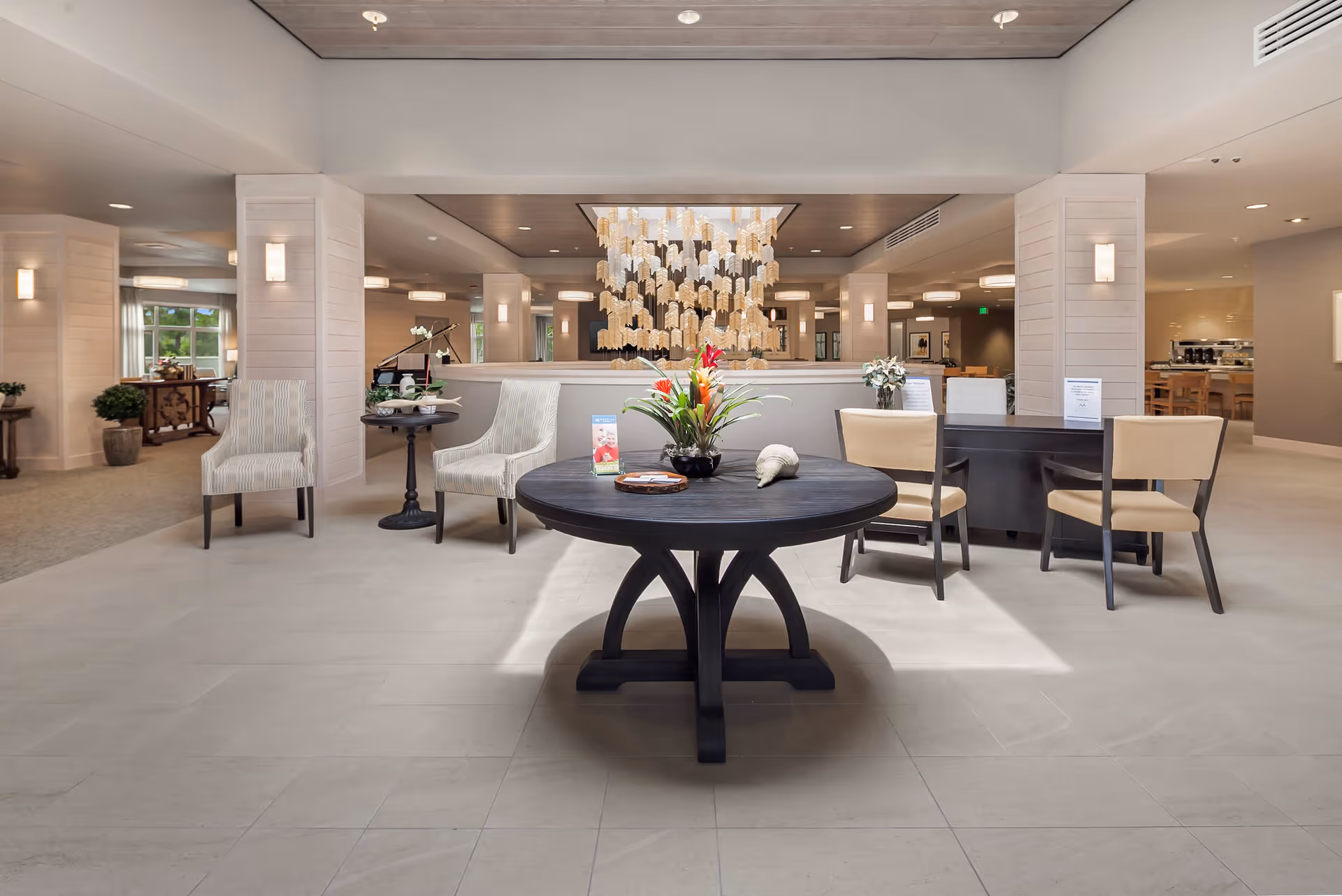 Spacious and well-lit senior living facility common area with a round black table in the center adorned with a floral arrangement and decorative items. Surrounding the table are several chairs and a small side table with a lamp. The background features a large chandelier, multiple columns, and additional seating areas with soft lighting.