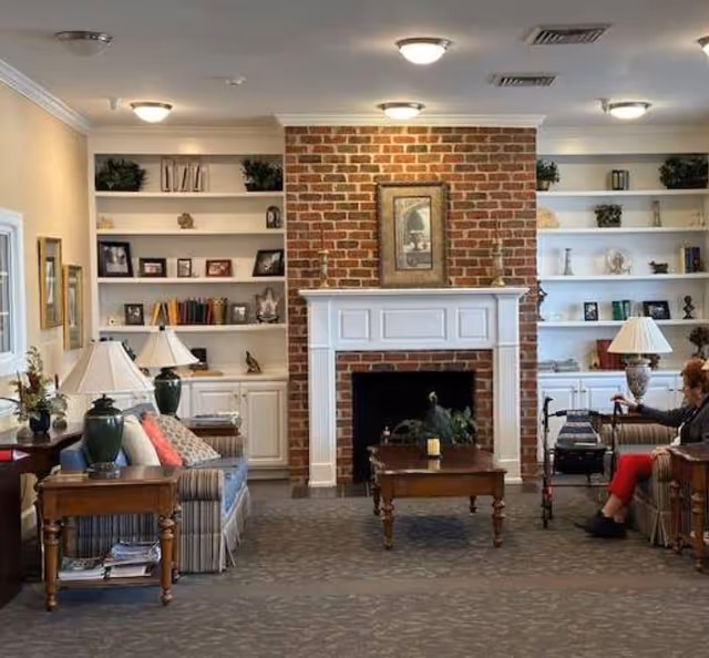 A cozy living room with a brick fireplace in the center, flanked by white built-in shelves filled with books, plants, and decorative items. There are two striped sofas facing each other with wooden side tables and lamps. A woman with a walker is seated on the right sofa. The room is softly lit with ceiling lights.