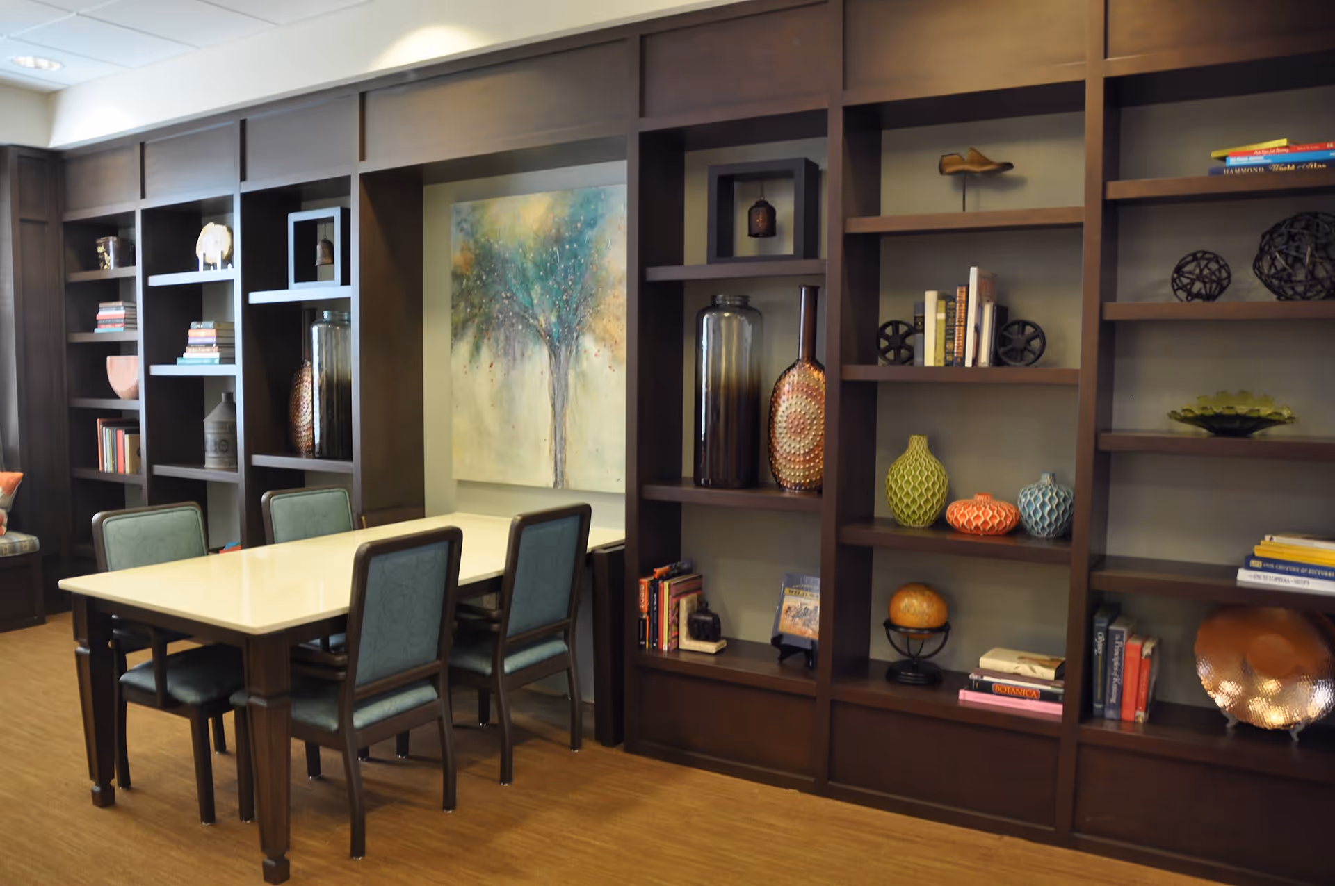 A room with a rectangular table surrounded by six chairs with green upholstery. Behind the table is a large built-in dark wood bookshelf filled with decorative items, books, and vases. A colorful painting of a tree is mounted on the wall in the center of the bookshelf. The floor has a wood-like finish.