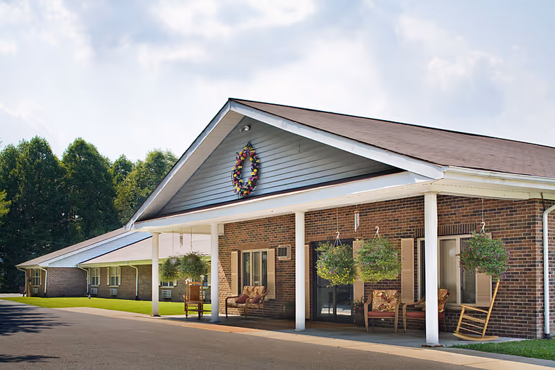Exterior view of a single-story brick building with a covered porch featuring hanging flower baskets and outdoor seating. The building has a gabled roof with a decorative wreath on the front. Trees and a clear sky are visible in the background.