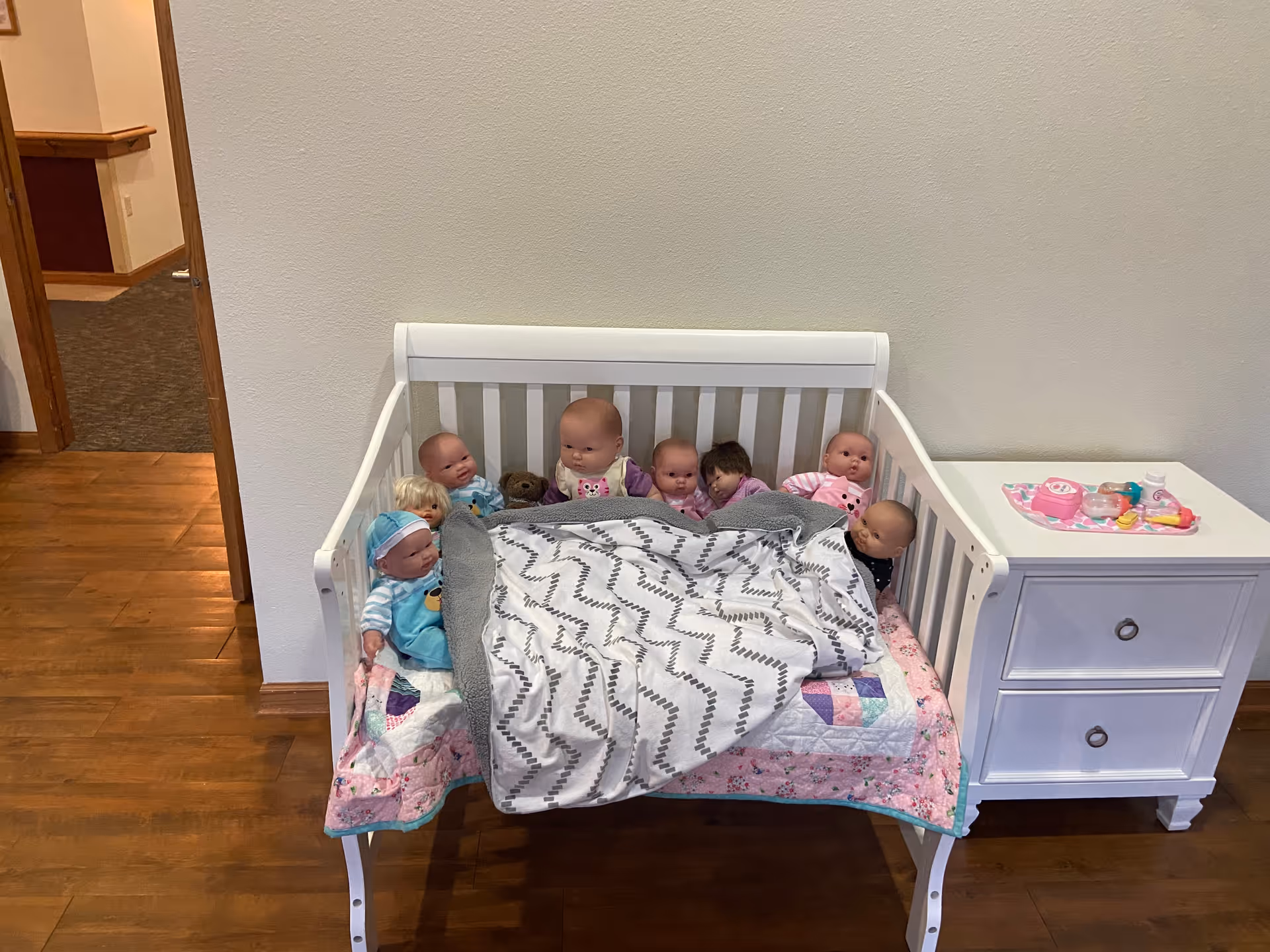 A small white crib with a patchwork quilt and a gray patterned blanket inside. The crib is filled with several baby dolls arranged sitting up, some partially covered by the blanket. Next to the crib is a white nightstand with two drawers, on top of which are toy baby bottles and a pink toy phone. The setting appears to be indoors with wooden flooring and a plain light-colored wall behind.