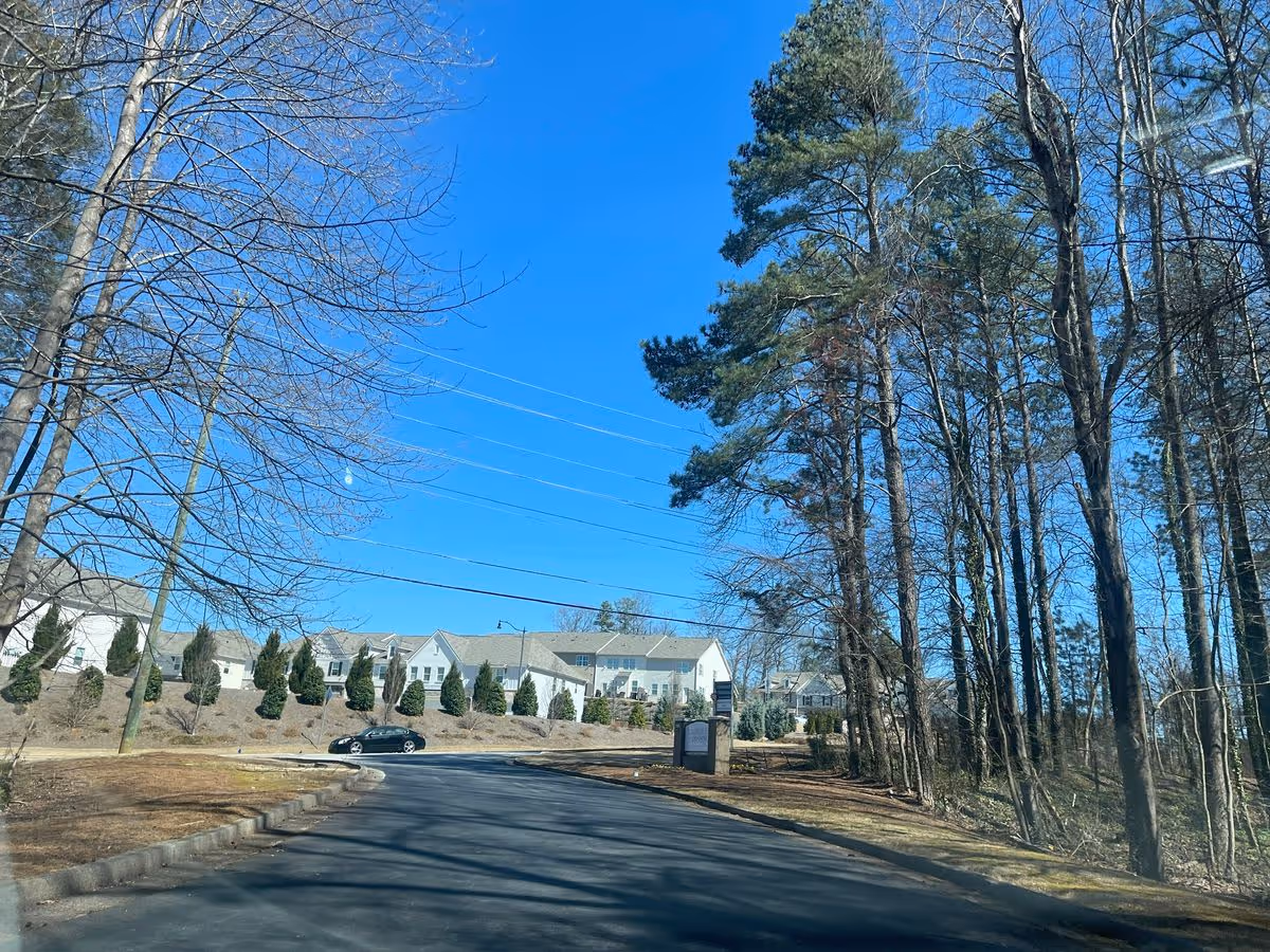 Paved driveway and trees leading to a cluster of white townhouse-style buildings under a clear blue sky.