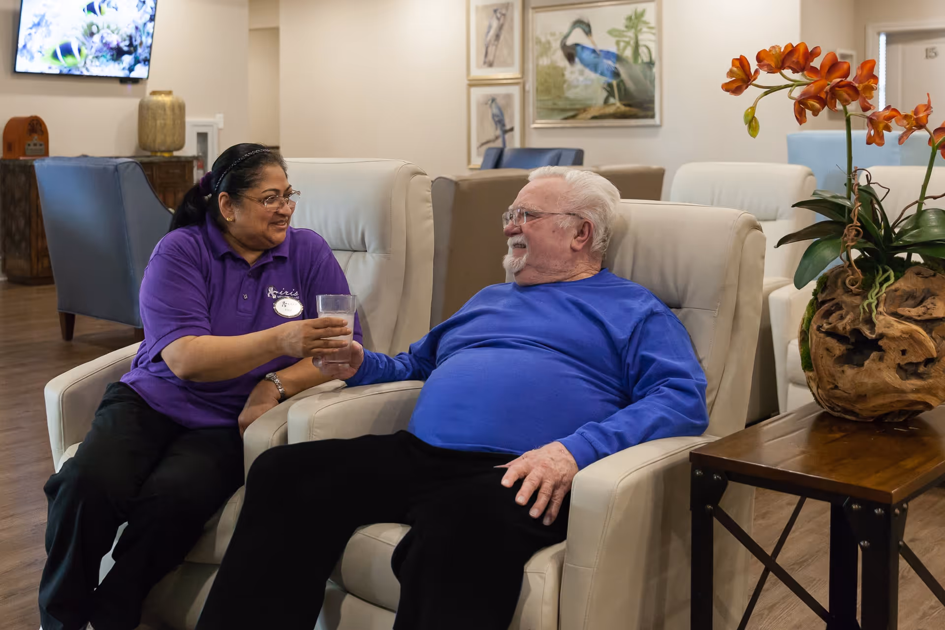 A caregiver in a purple shirt with an Iris Memory Care of Edmond badge is sitting next to an elderly man in a blue shirt on a beige armchair. They are smiling at each other while the caregiver hands the man a glass of water. The room has comfortable seating, framed bird artwork on the wall, and a wooden table with an orange orchid plant.