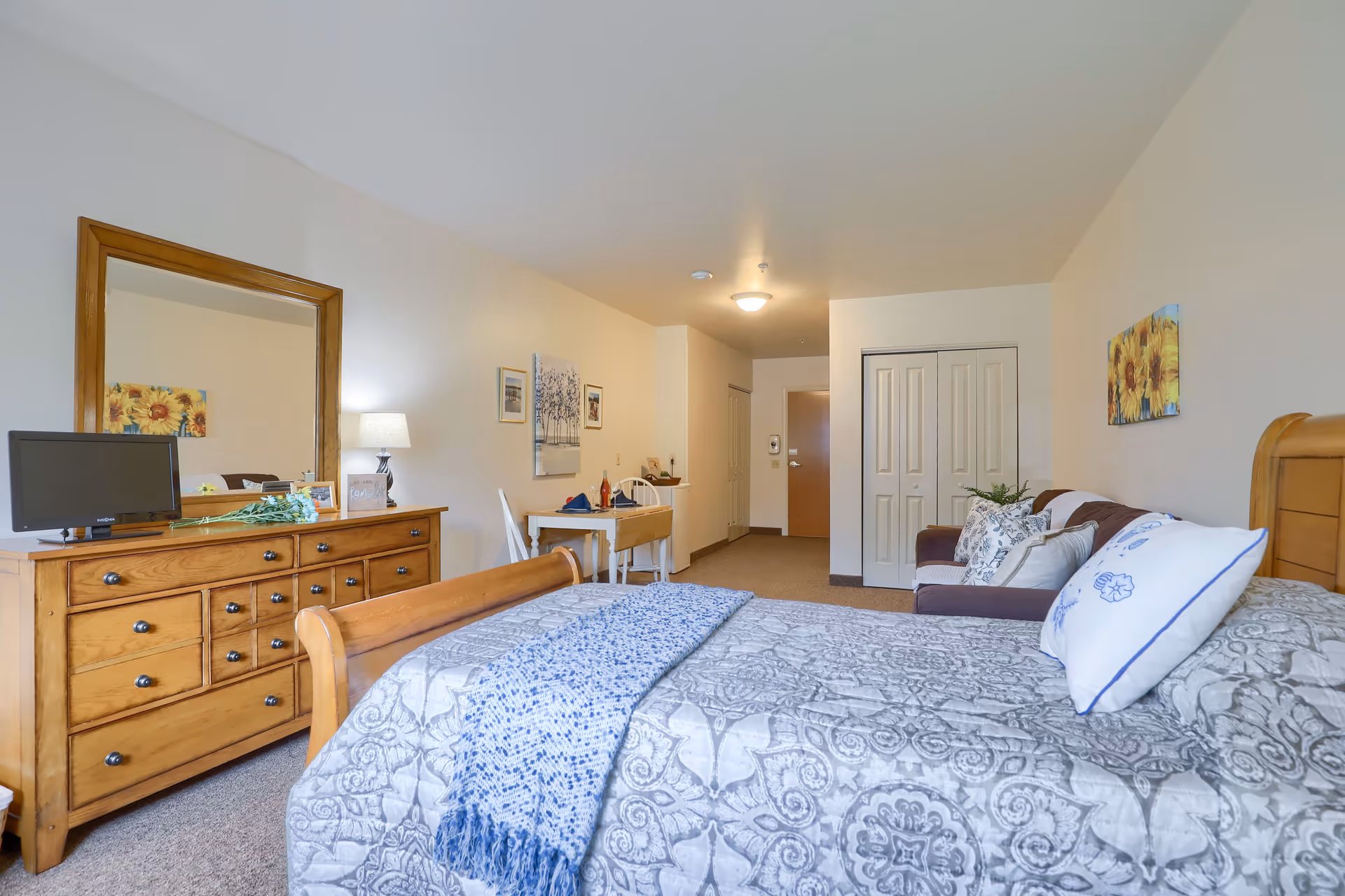 A well-lit senior living bedroom featuring a wooden bed with patterned bedding and pillows, a wooden dresser with a mirror and a small TV on top, a small table with two chairs, a brown sofa with decorative pillows, and wall art including sunflower paintings.
