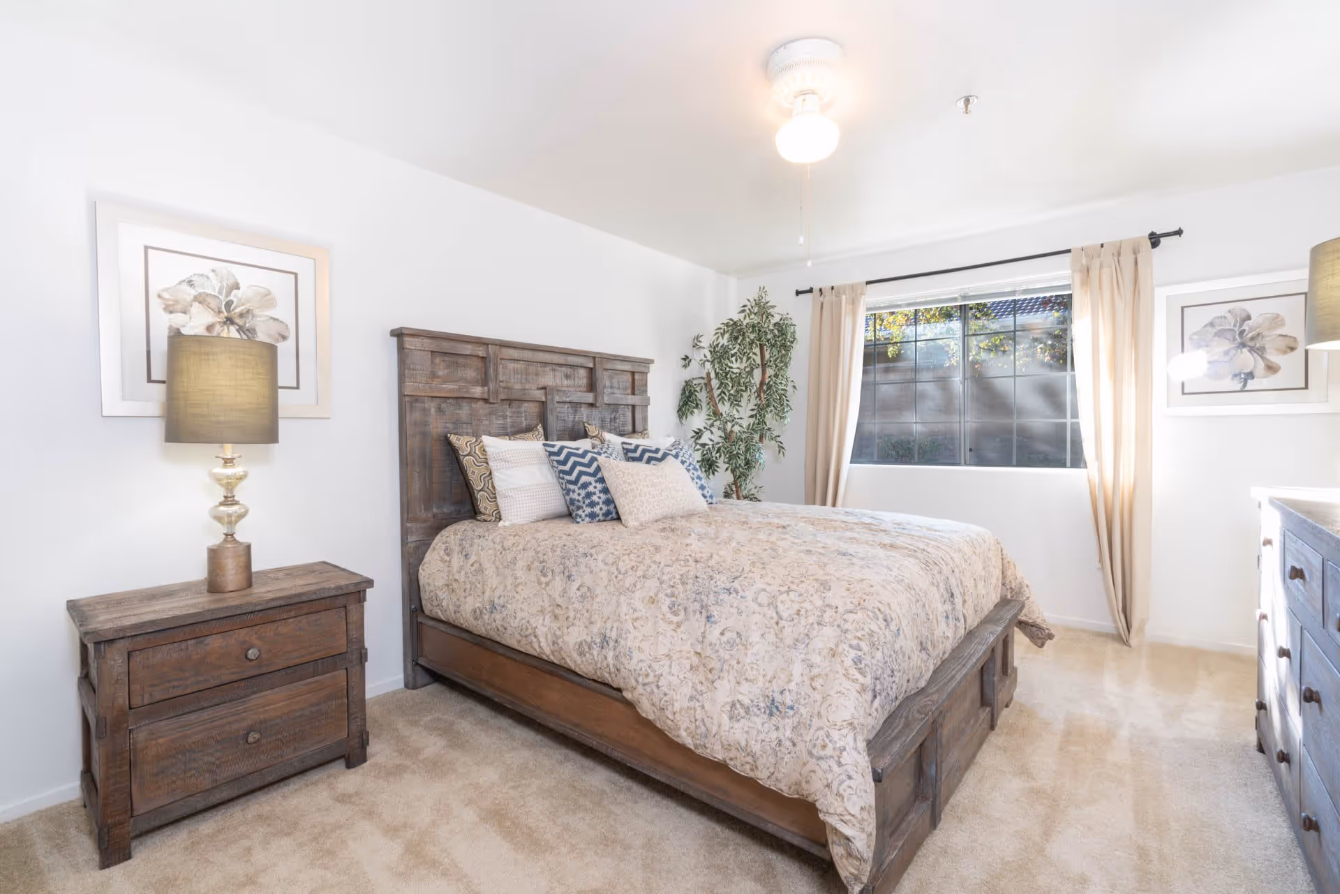 A bright bedroom with a large wooden bed featuring a patterned bedspread and multiple decorative pillows. There is a wooden nightstand with a lamp and a framed flower picture on the wall to the left of the bed. A large window with beige curtains lets in natural light, and there is a tall green plant in the corner. On the right side, there is a wooden dresser with another lamp and a framed flower picture above it. The room has light-colored carpet and white walls.