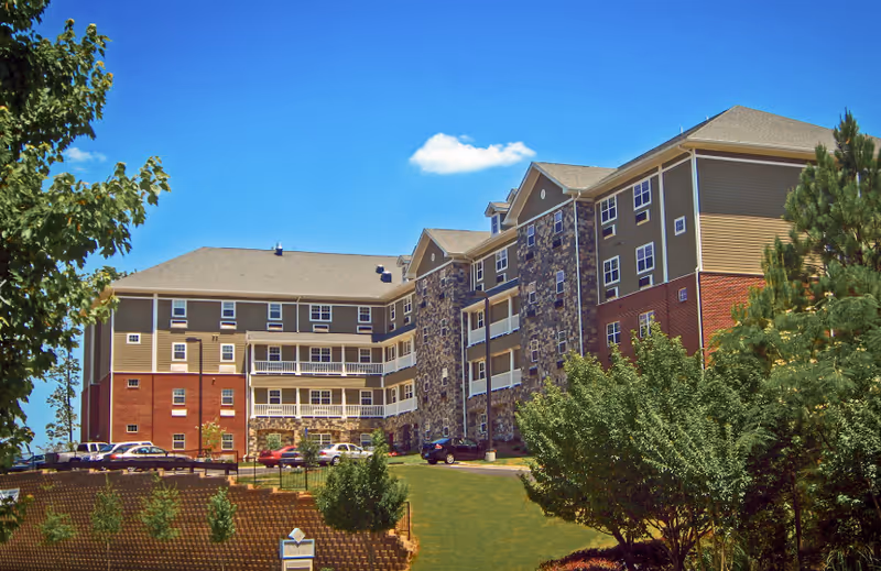 Exterior view of a multi-story senior living facility building with a combination of brick, stone, and siding. The building is surrounded by green trees and a well-maintained lawn under a clear blue sky with a few clouds. Several cars are parked near the building.