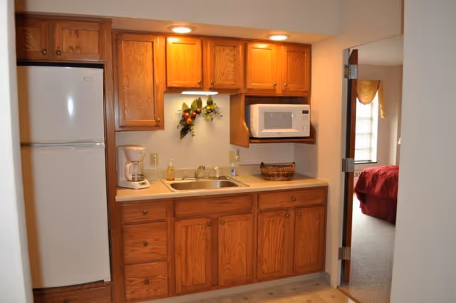 Small kitchenette with oak cabinets, a sink, white refrigerator and microwave, and an open doorway into a bedroom.