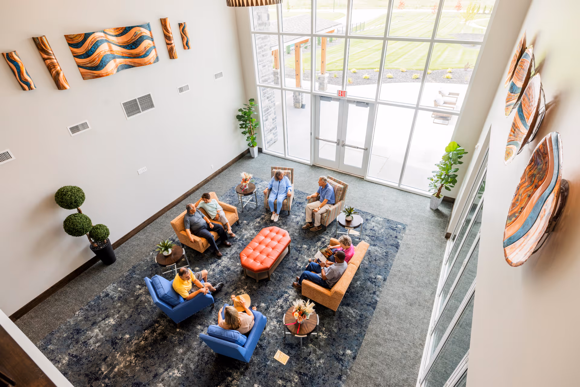 A bright and spacious living room area with large floor-to-ceiling windows letting in natural light. The room features a blue patterned rug, two blue armchairs, two orange sofas, a red tufted ottoman in the center, and several small round tables with plants and floral arrangements. There are seven elderly people sitting and conversing comfortably. The walls are decorated with abstract wooden art pieces and there are two potted plants near the windows.