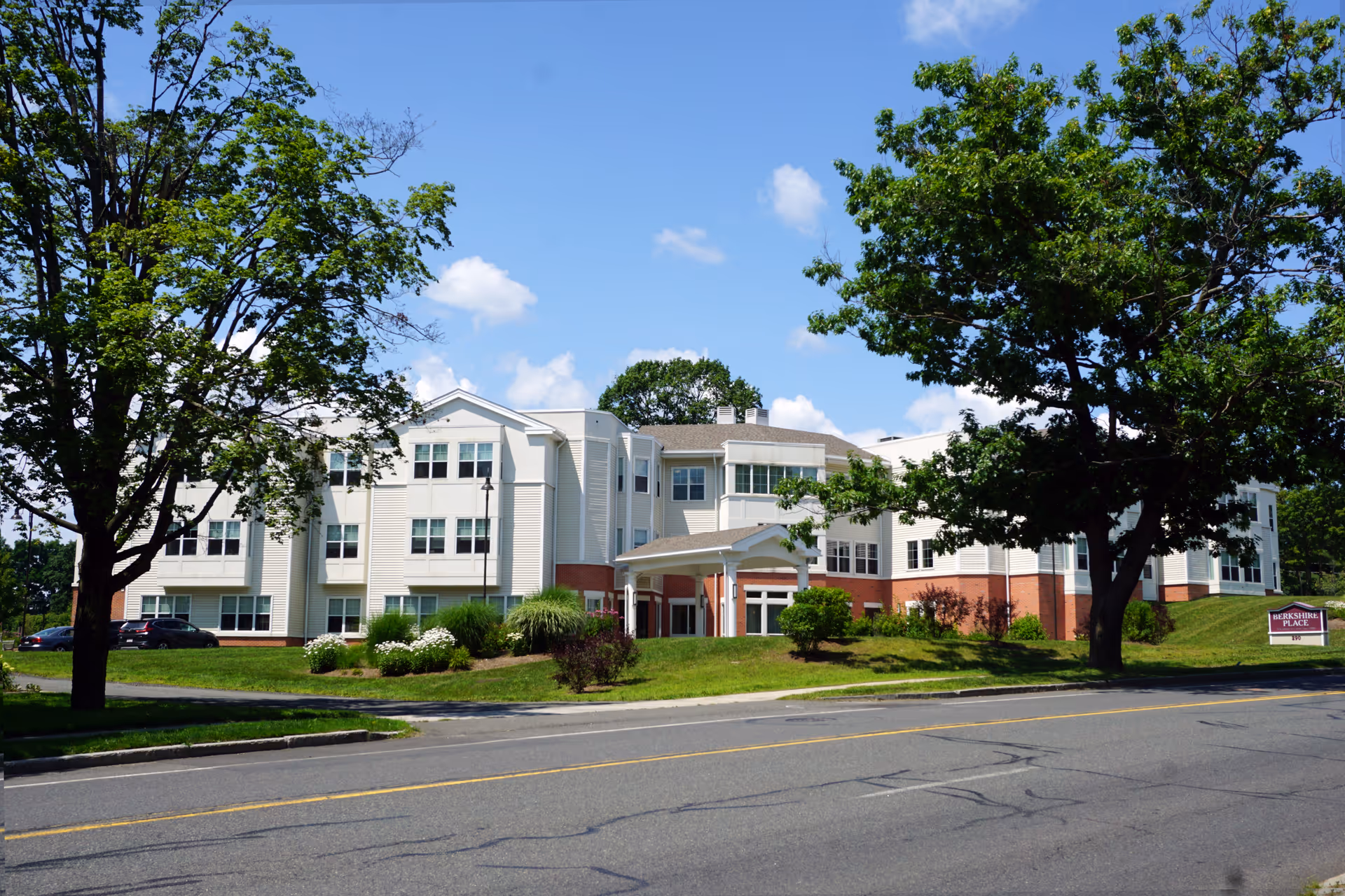 Exterior view of a multi-story senior living facility named Berkshire Place, surrounded by green trees and landscaping under a blue sky with some clouds.