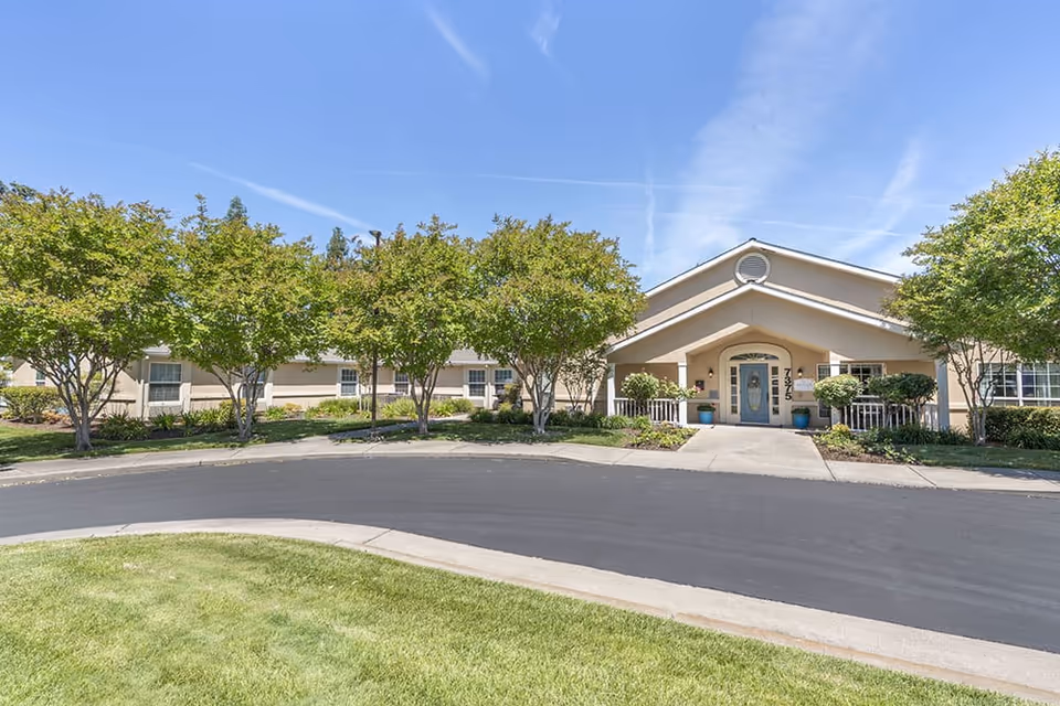 Front exterior view of Brookdale Sylvan Ranch facility showing a beige building with a peaked roof entrance, surrounded by green trees and landscaping under a clear blue sky.