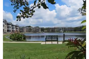 A peaceful outdoor scene at Sunnyside Village featuring a bench facing a large pond surrounded by green grass, plants, and multi-story residential buildings under a partly cloudy sky.