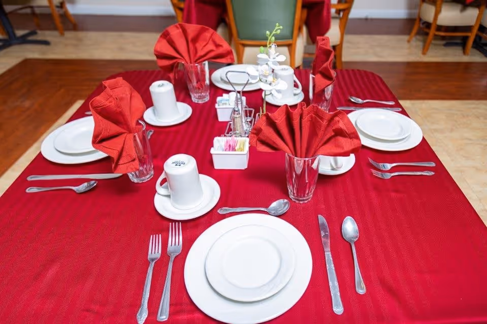 A dining table set for four with a red tablecloth, white plates, cups, silverware, and glasses with red folded napkins. There is a small white flower centerpiece and containers with sugar packets and salt and pepper shakers in the middle of the table.