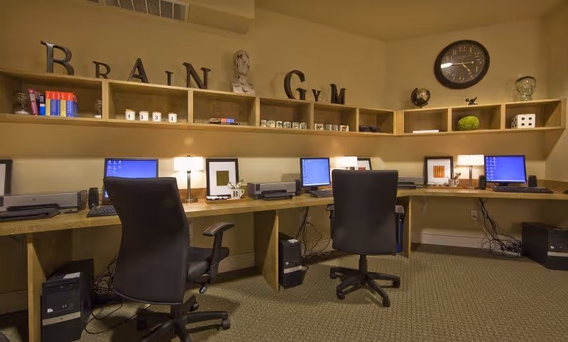 A room with a long wooden desk holding three computer workstations, each with a black office chair. Above the desk are wooden shelves containing books, decorative items, and large letters spelling out 'BRAIN GYM'. A round wall clock is mounted on the wall to the right.