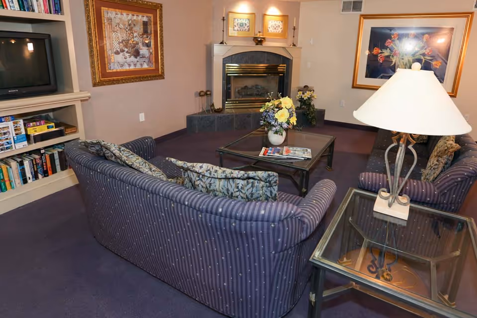 A cozy living room area with two striped sofas arranged around a glass coffee table holding a vase of yellow flowers and magazines. There is a fireplace with decorative items above it, framed artwork on the walls, a side table with a lamp, and a built-in bookshelf with books and games. The room has a purple carpet and soft lighting.