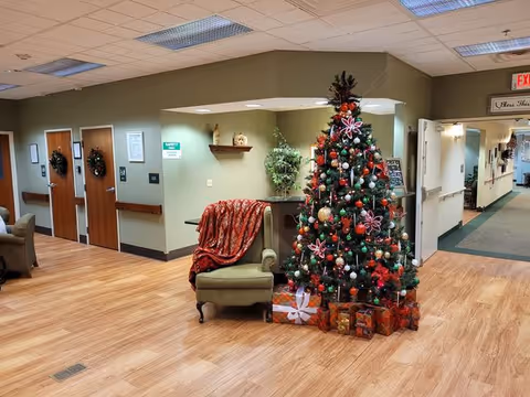 A decorated Christmas tree with ornaments and wrapped presents underneath stands next to a green armchair draped with a red blanket in a hallway of a senior living facility. The hallway has wooden floors, beige walls, and several doors adorned with wreaths. There are plants and shelves with decorative items on the walls.