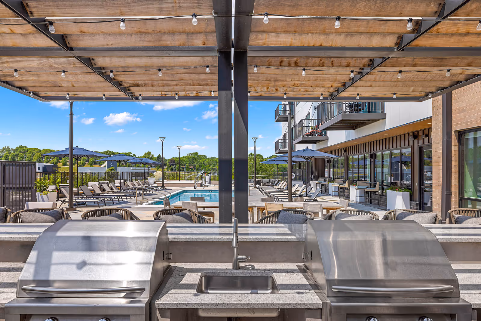 Outdoor patio area at Avidor Omaha featuring two stainless steel grills with a sink in between under a wooden pergola, overlooking a swimming pool surrounded by lounge chairs and umbrellas, with a multi-story building and balconies in the background under a clear blue sky.