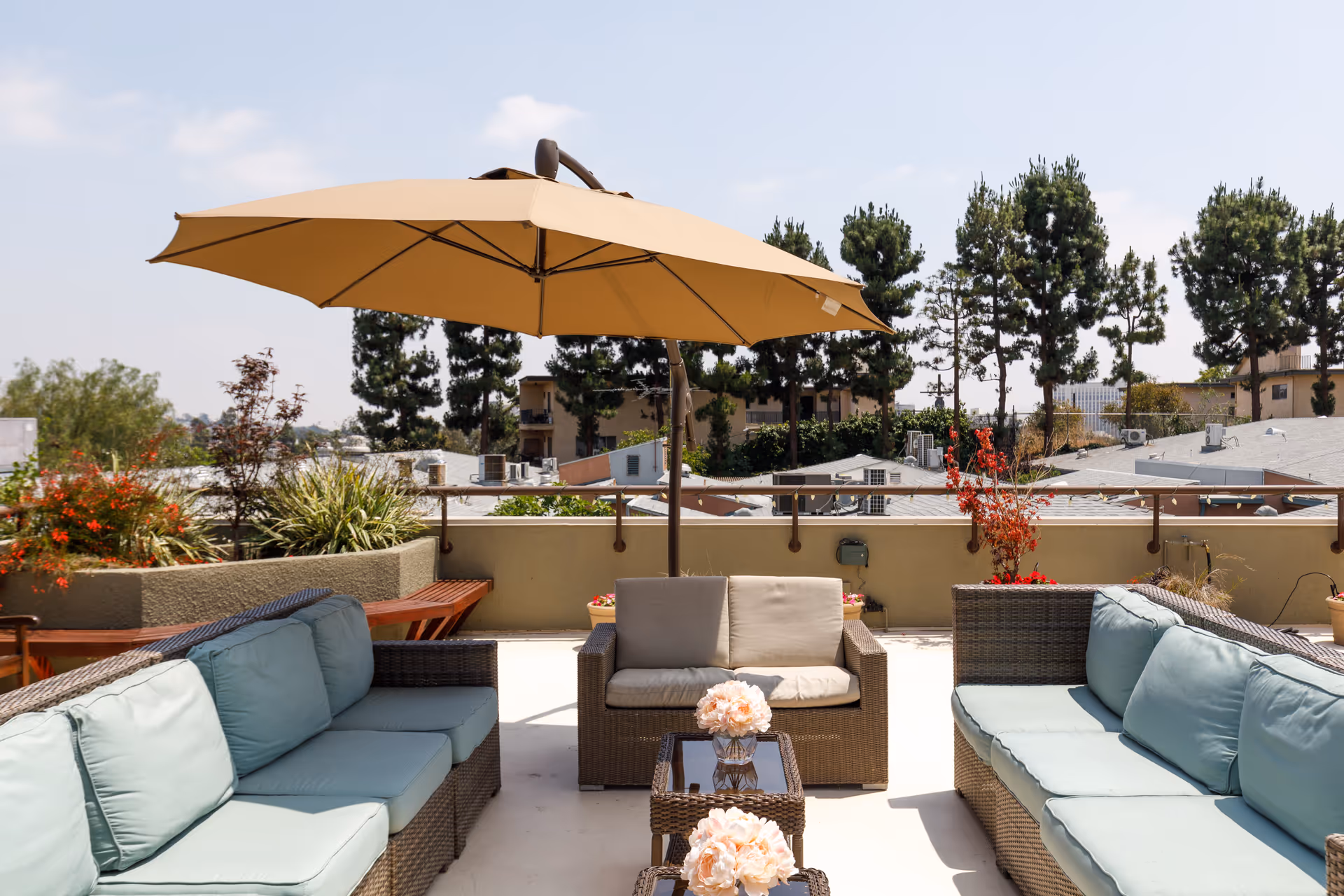 Rooftop patio with wicker sofas, a central umbrella, small tables with flowers, and potted plants overlooking nearby rooftops and trees.