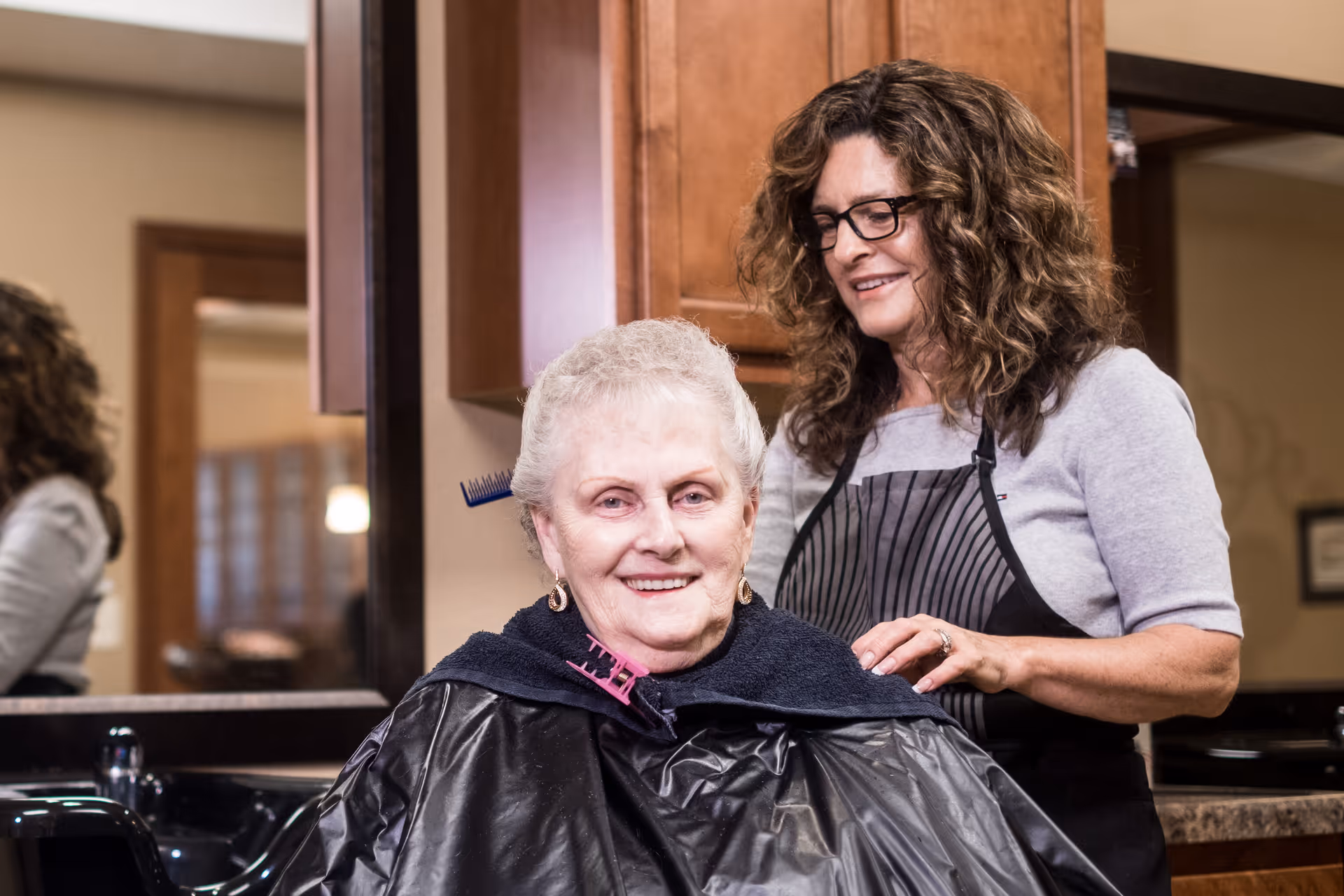An elderly woman with short white hair is sitting in a salon chair wearing a black cape, smiling while a hairstylist with curly hair and glasses stands behind her, preparing to style her hair in a well-lit salon setting.