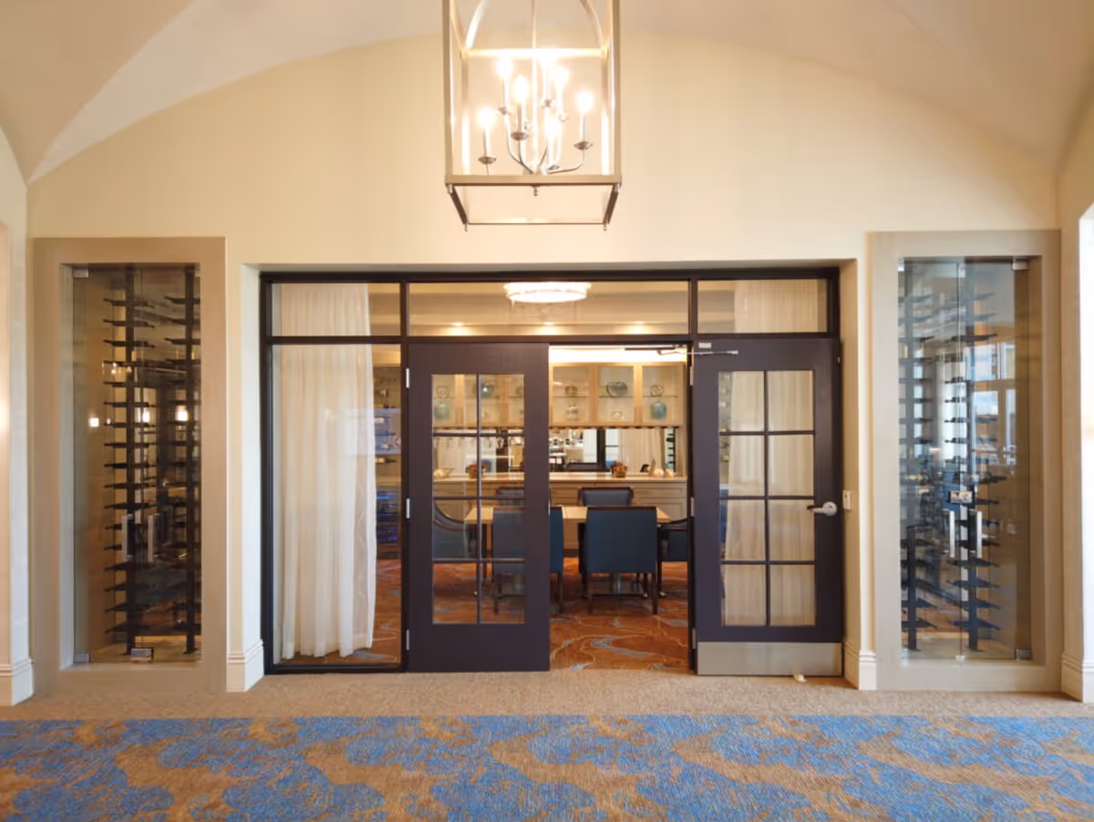 Entrance to a dining room with double glass doors framed in dark wood. On either side of the doors are glass-enclosed wine racks. The room has a patterned carpet and a chandelier hanging from a vaulted ceiling.