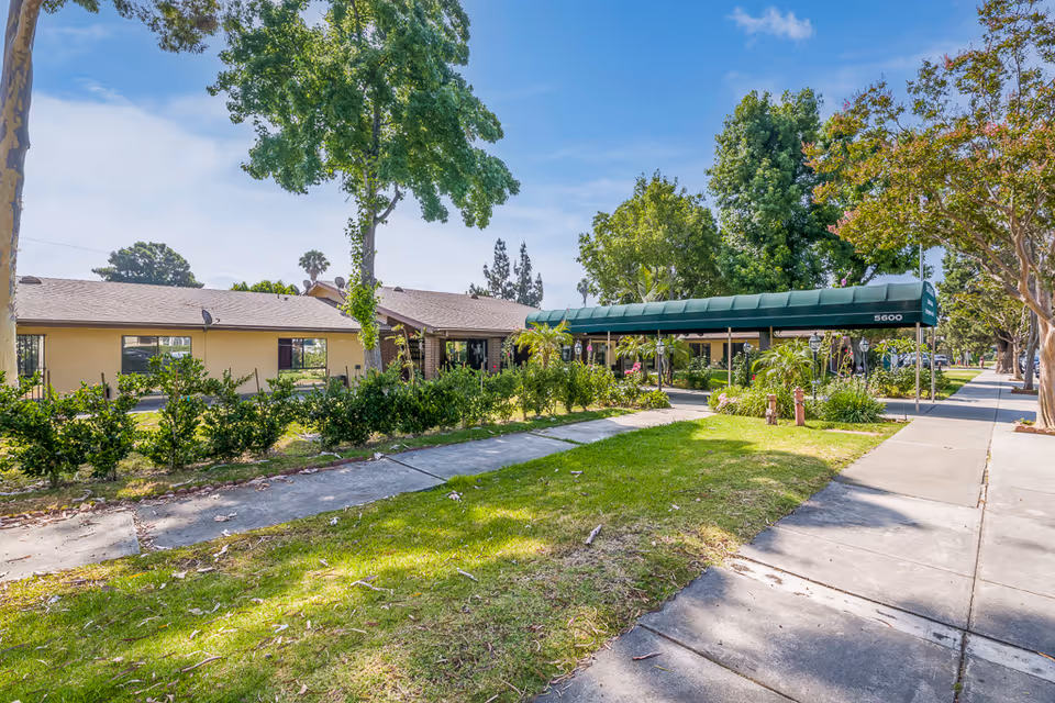 Exterior view of Santa Anita Assisted Living facility showing a single-story building with a green awning over the entrance, surrounded by trees, bushes, and a sidewalk under a clear blue sky.