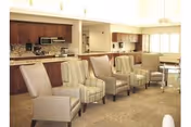 Communal seating area with a row of armchairs facing a kitchenette and dining tables in the background.