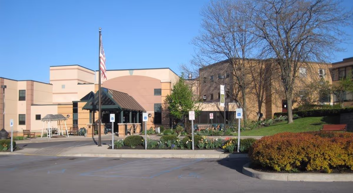 Exterior view of The Inn at Menorah Park building with an American flag on a flagpole in front, surrounded by landscaped bushes, trees, and flower beds. There are several handicap parking spaces and benches near the entrance under a covered walkway.