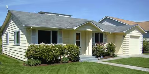 Front exterior of a single-story yellow house with shrubs, lawn, and a driveway.