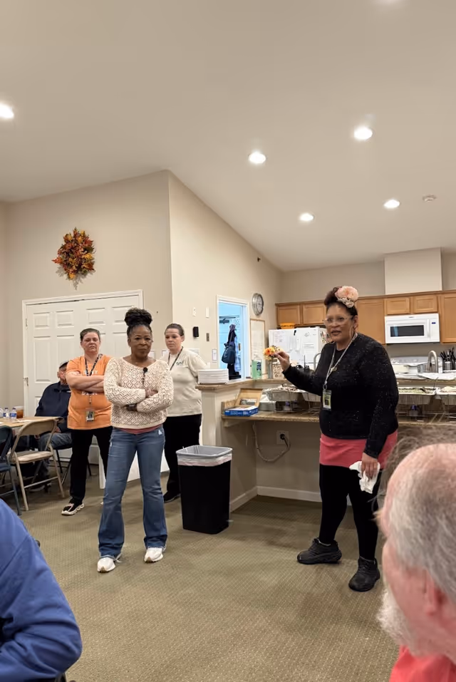 A group of four women standing in a room with a kitchen area in the background. One woman is speaking and gesturing with her hand, while the others listen. There are chairs and people seated partially visible in the foreground. The room has beige walls, a clock on the wall, and overhead recessed lighting.