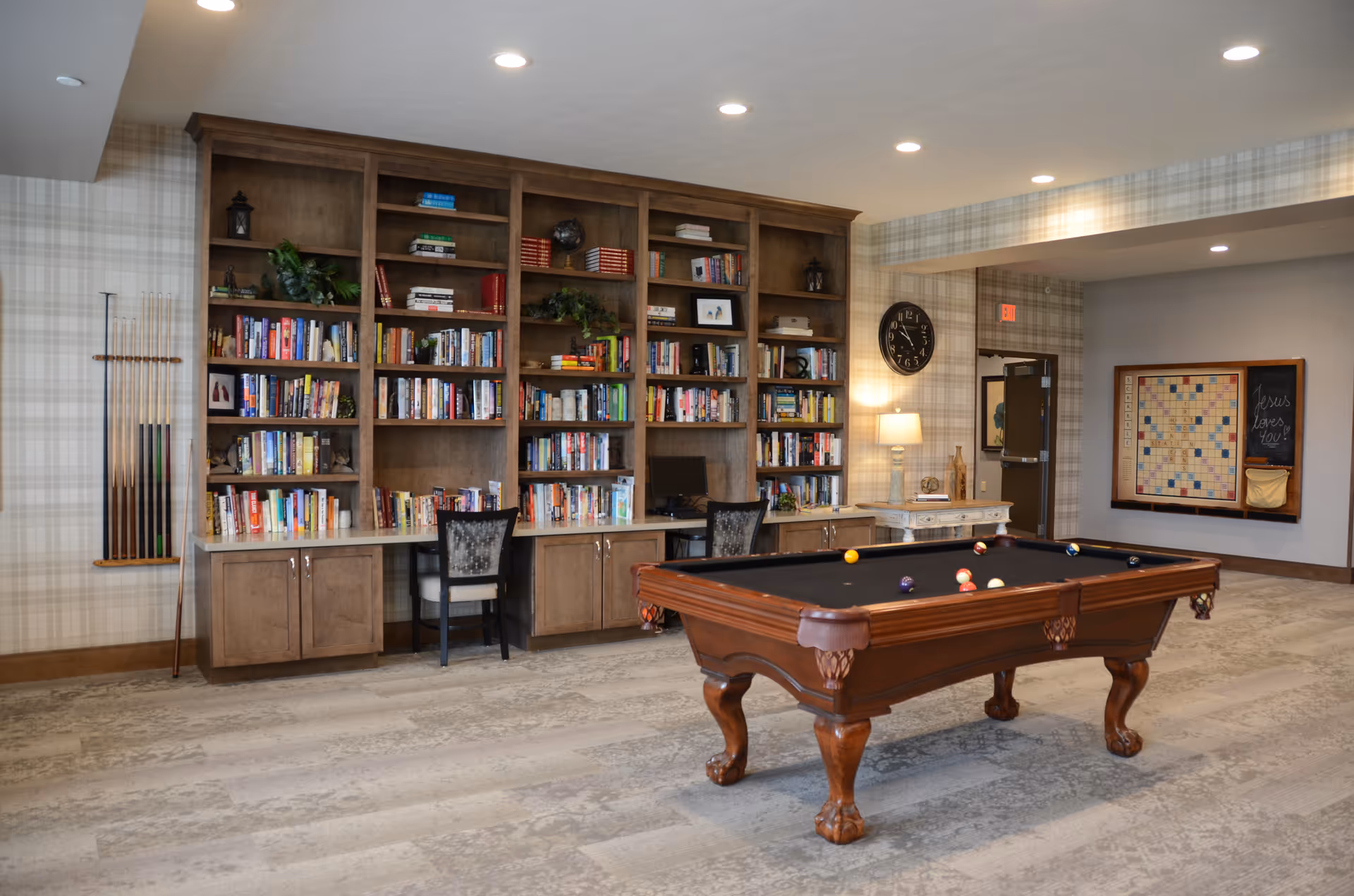 A recreational room featuring a wooden pool table with billiard balls arranged on it, a large wooden bookshelf filled with books and decorative items, two chairs at a built-in desk area, a wall-mounted rack holding pool cues, a clock on the wall, a lamp on a side table, and a framed Scrabble board with a chalkboard that reads 'Jesus loves you!'