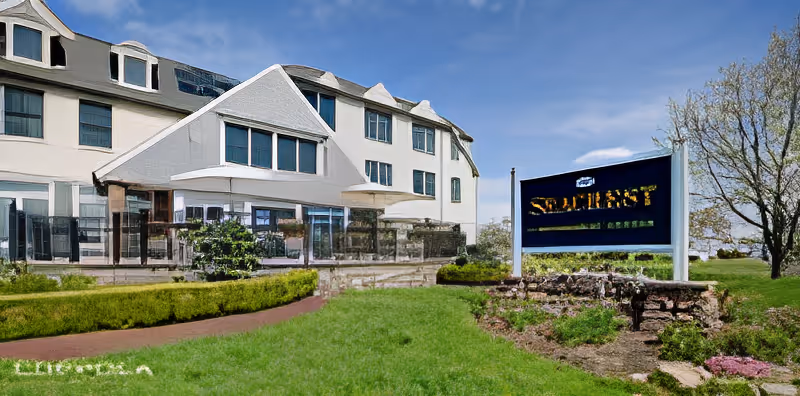 Exterior view of a multi-story assisted living facility building with large windows and a well-maintained lawn and garden area in front. There is a prominent sign on the right side of the image displaying the name 'Seacrest Assisted Living & Memory Care'. Trees and a clear blue sky are visible in the background.