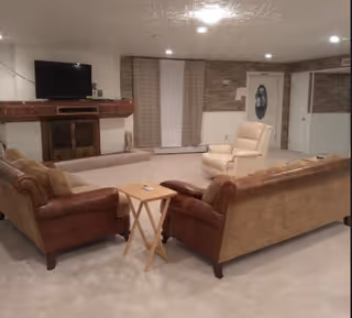Basement living room with two sofas and a recliner facing a wall-mounted TV above a mantel, neutral carpeting and paneled walls.