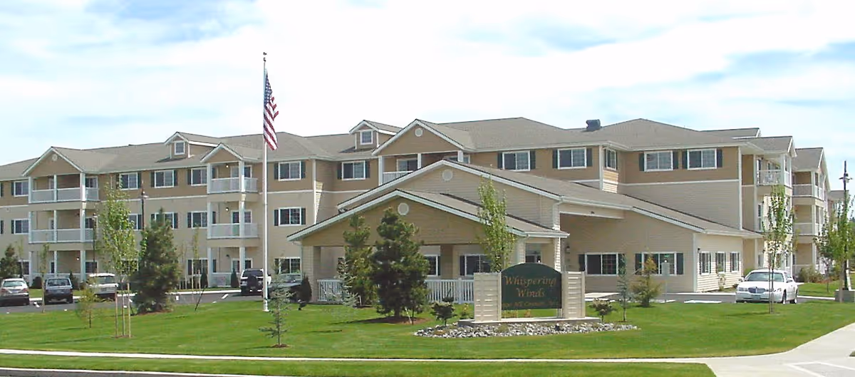 Exterior view of Whispering Wind Retirement facility showing a large, multi-story beige building with balconies, a green lawn with small trees, an American flag on a flagpole, and a sign with the facility's name in front.