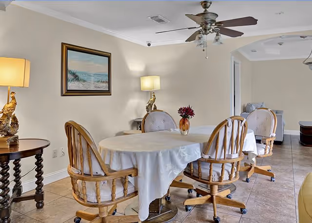 A dining area with a rectangular table covered with a white tablecloth, surrounded by four wooden chairs with cushioned seats and backs on wheels. The room has beige walls, a ceiling fan with lights, two table lamps with decorative bases, a framed painting of a beach scene on the wall, and a tiled floor. In the background, there is an archway leading to another room with a sofa.
