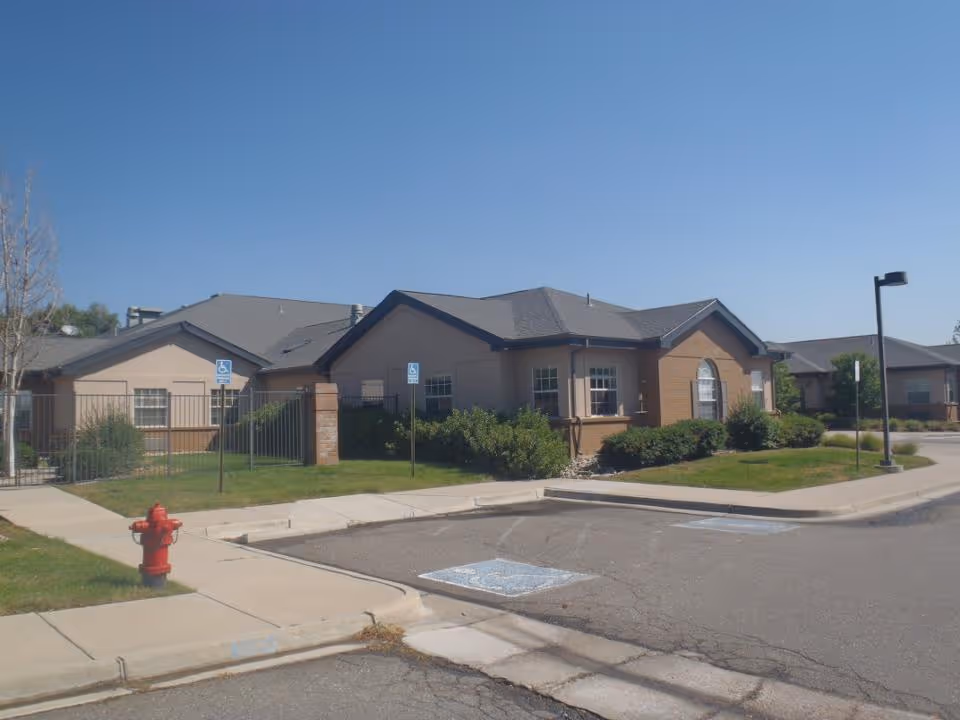 Exterior view of a single-story senior living facility building with beige and brick walls, surrounded by green bushes and a lawn. There are two handicap parking spaces and a red fire hydrant near the sidewalk under a clear blue sky.