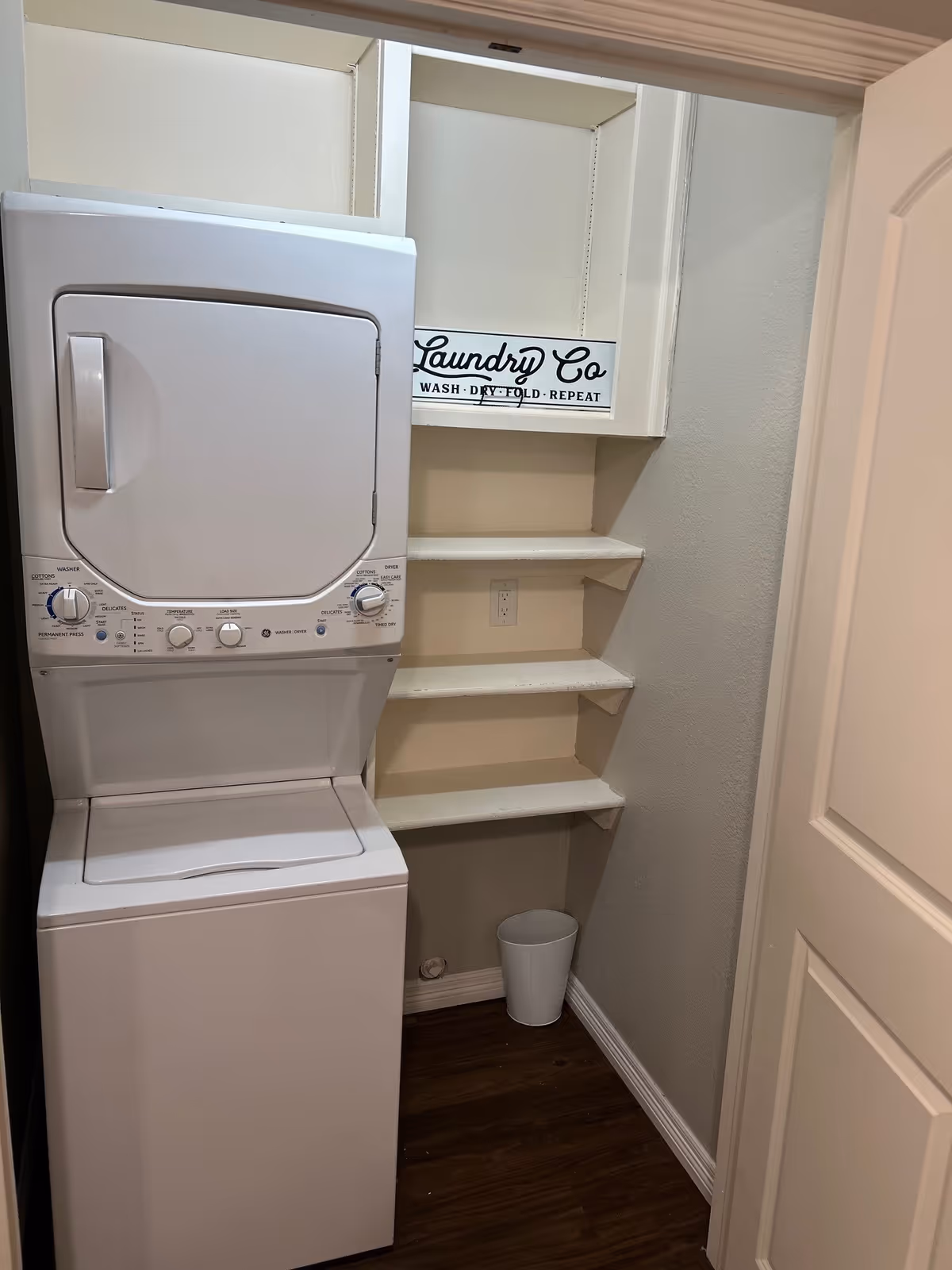 Stacked white washer and dryer in a small laundry closet with built-in shelves and a 'Laundry Co' sign.