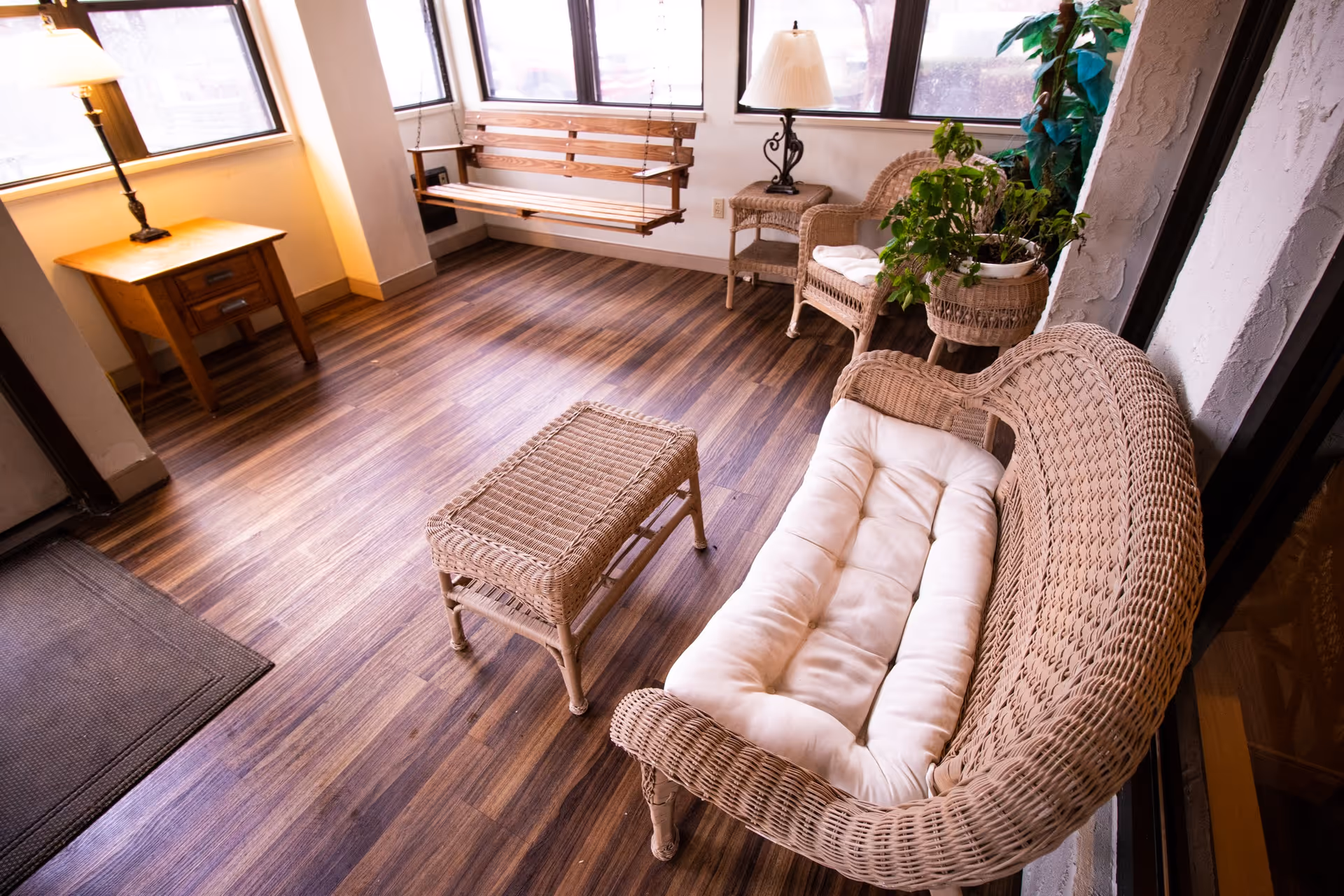 A cozy indoor sitting area with wicker furniture including a cushioned loveseat, a wicker coffee table, and a wicker chair with a lamp on a side table. There is a wooden swing bench hanging near the windows and several green plants adding a touch of nature to the space. The floor is made of wood with a warm tone.