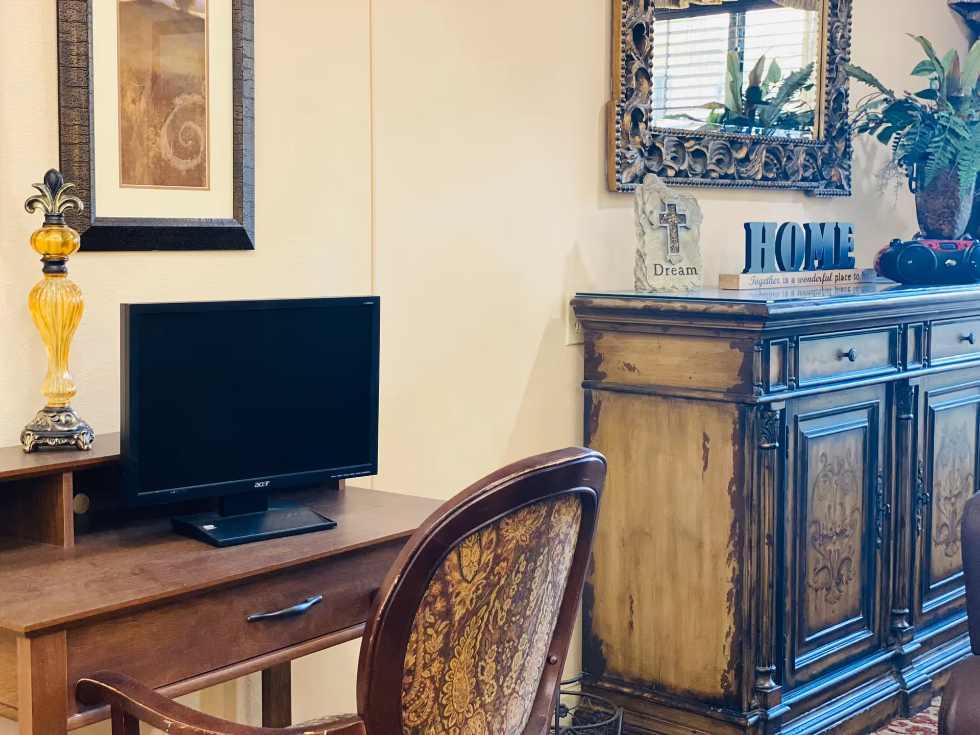 A cozy interior corner featuring a wooden desk with a black Acer monitor, an ornate yellow glass lamp, and a framed artwork on the wall. Next to the desk is a vintage-style wooden cabinet with decorative carvings, topped with a mirror, a stone plaque with a cross and the word 'Dream', a decorative 'HOME' sign, and a potted plant.