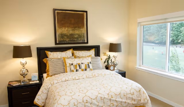 A neatly made bed with patterned white and yellow bedding and multiple pillows in a bedroom. There are two bedside tables with matching lamps, a small plant, and a clock. A large window shows a view of a green outdoor area with people in the distance. A framed abstract painting hangs above the bed.