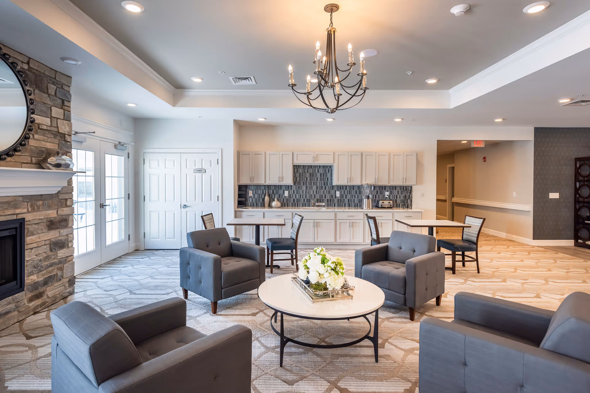 A bright and spacious common area in a senior living facility featuring four gray armchairs arranged around a round white coffee table with a floral centerpiece. The room has a stone fireplace on the left, a chandelier hanging from the ceiling, and a kitchenette with white cabinets and a tiled backsplash in the background. There are also two small dining tables with chairs near the kitchenette and double glass doors letting in natural light.