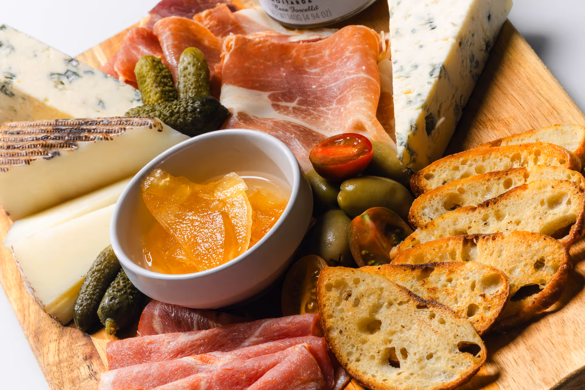 A wooden platter with an assortment of charcuterie including slices of cured meats, wedges of cheese, pickles, green olives, cherry tomato halves, toasted bread slices, and a small bowl of orange marmalade or jelly.