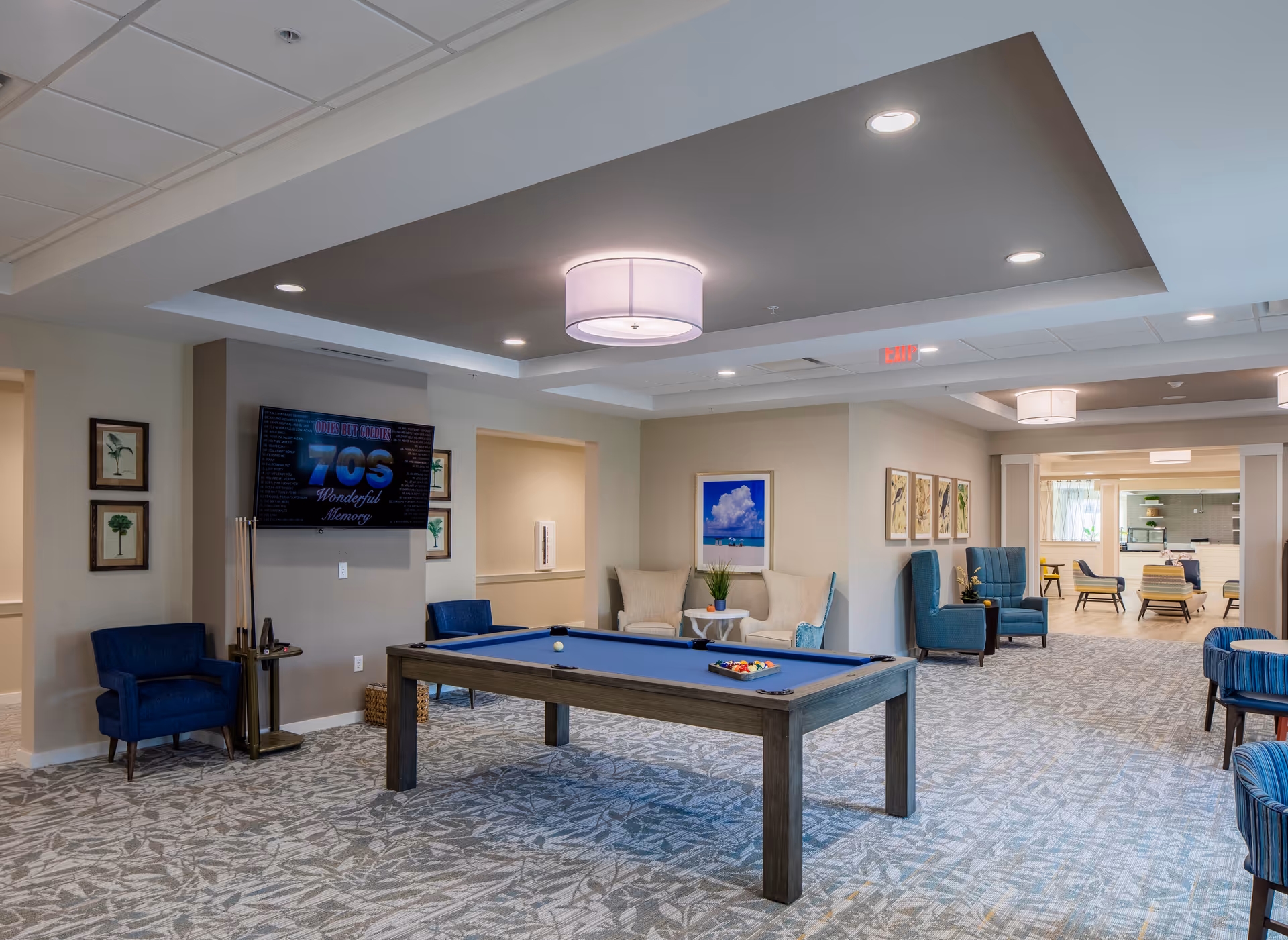 A spacious and well-lit common area in a senior living facility featuring a pool table with balls arranged on it. The room has comfortable seating including blue and beige armchairs, framed artwork on the walls, and a flat-screen TV mounted on a wall displaying '70s Wonderful Memory'. The ceiling has recessed lighting and a modern light fixture. In the background, there is another seating area with more chairs and tables.