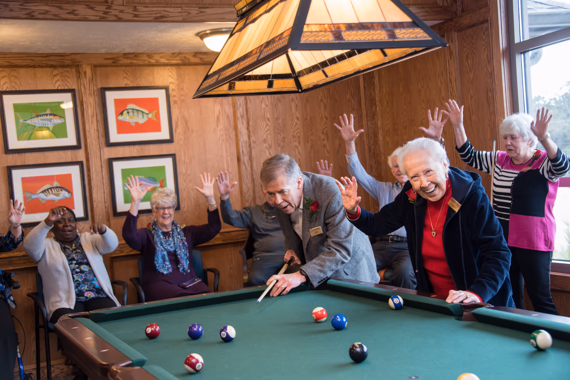 A group of elderly people enjoying a game of pool in a wood-paneled room. One man is taking a shot while others cheer and raise their hands in excitement. The room has framed fish artwork on the wall and a large stained glass light fixture above the pool table.